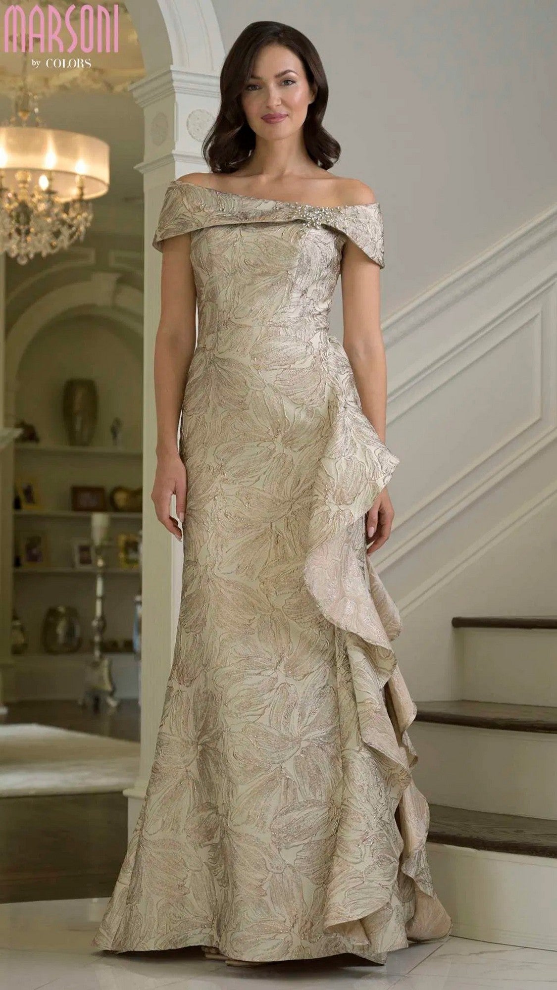 A woman stands indoors on a staircase wearing the Marsoni MV1381 long formal dress in champagne, featuring metallic brocade fabric, an off-the-shoulder neckline, and elegant ruffle details.
